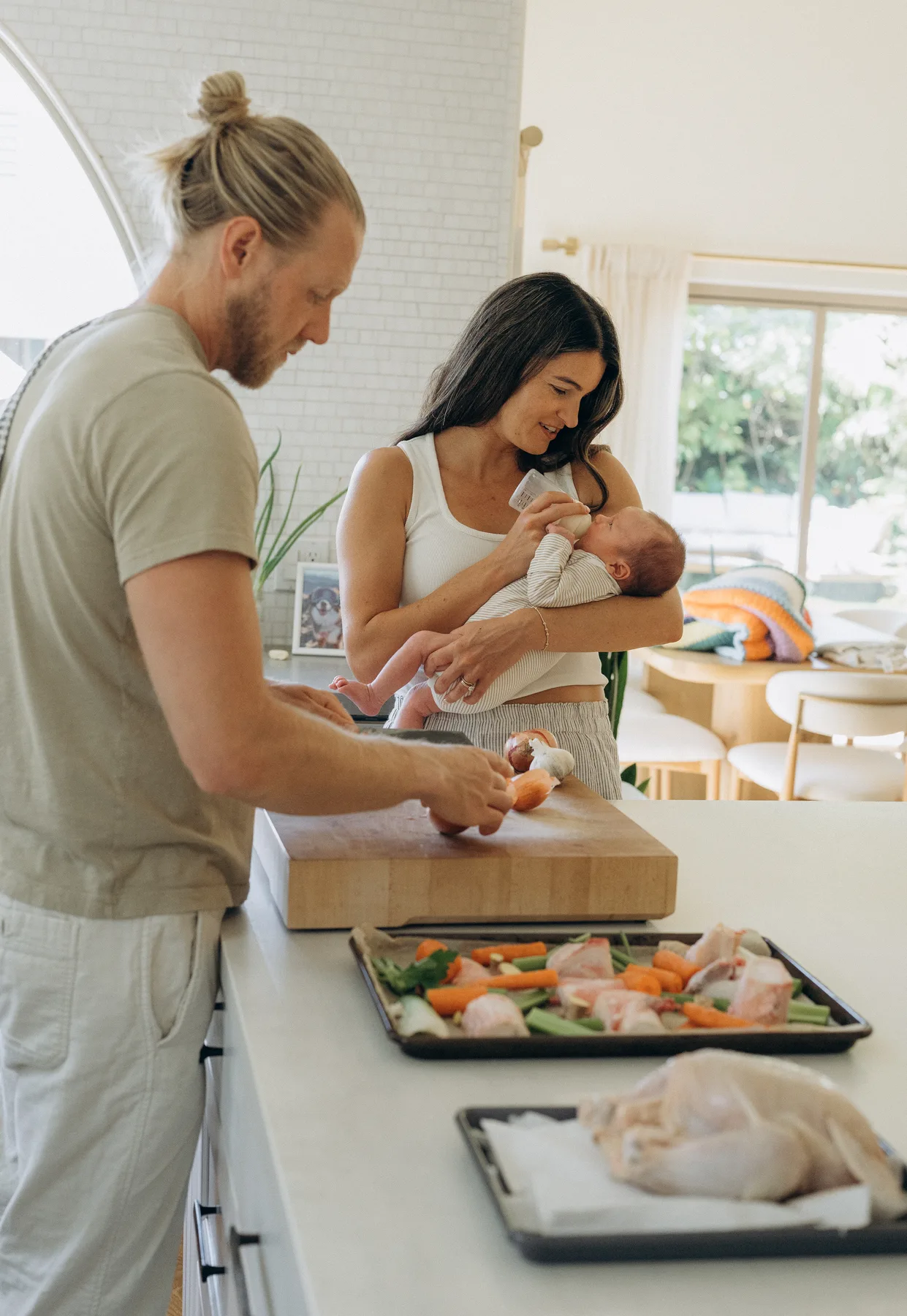 Adam prepares vegetables while his wife holds their baby in the kitchen.